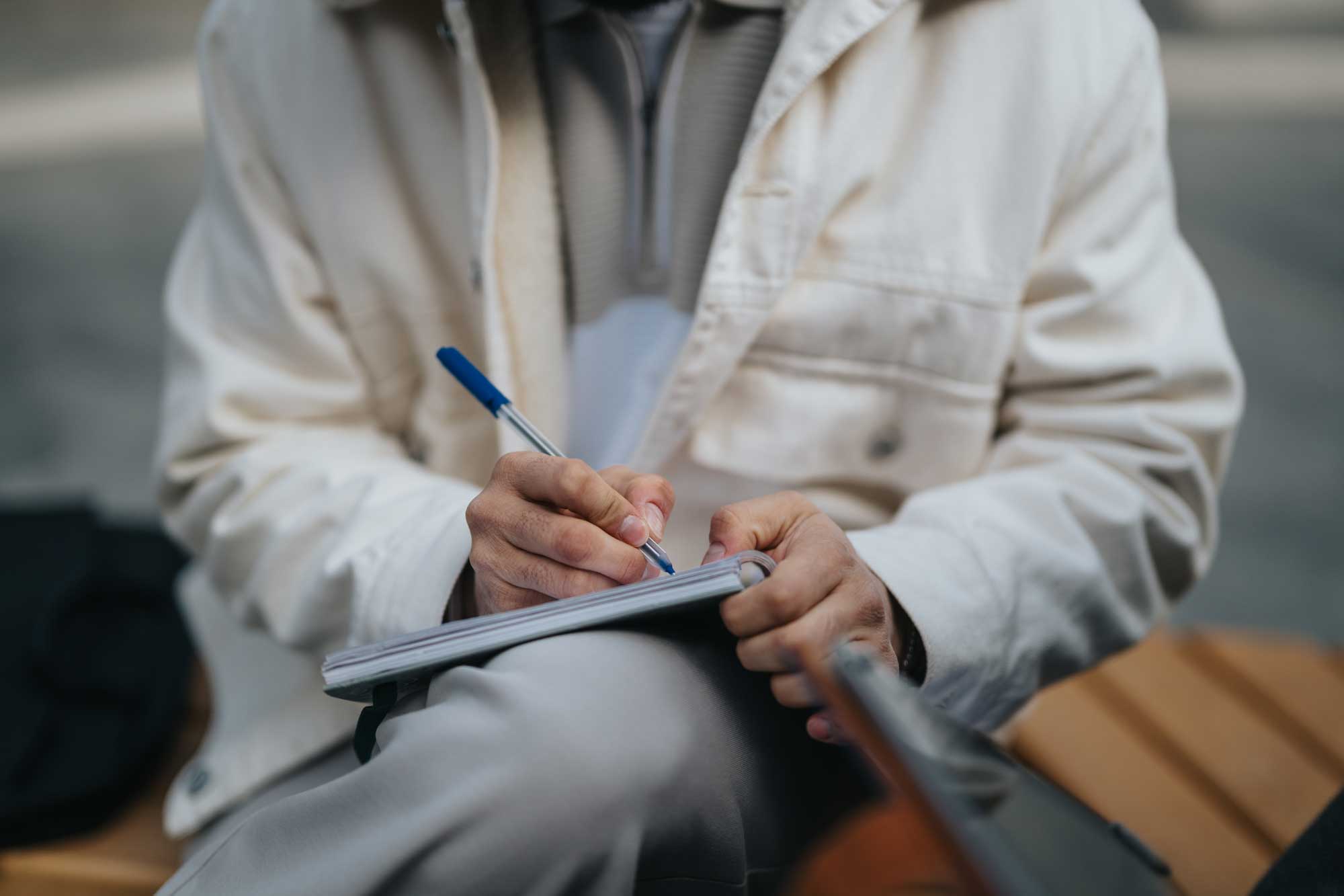 A person dressed in a light-colored jacket is sitting outside and writing in a notebook using a blue-ink pen. The scene suggests casual learning, journaling, or expressing thoughts